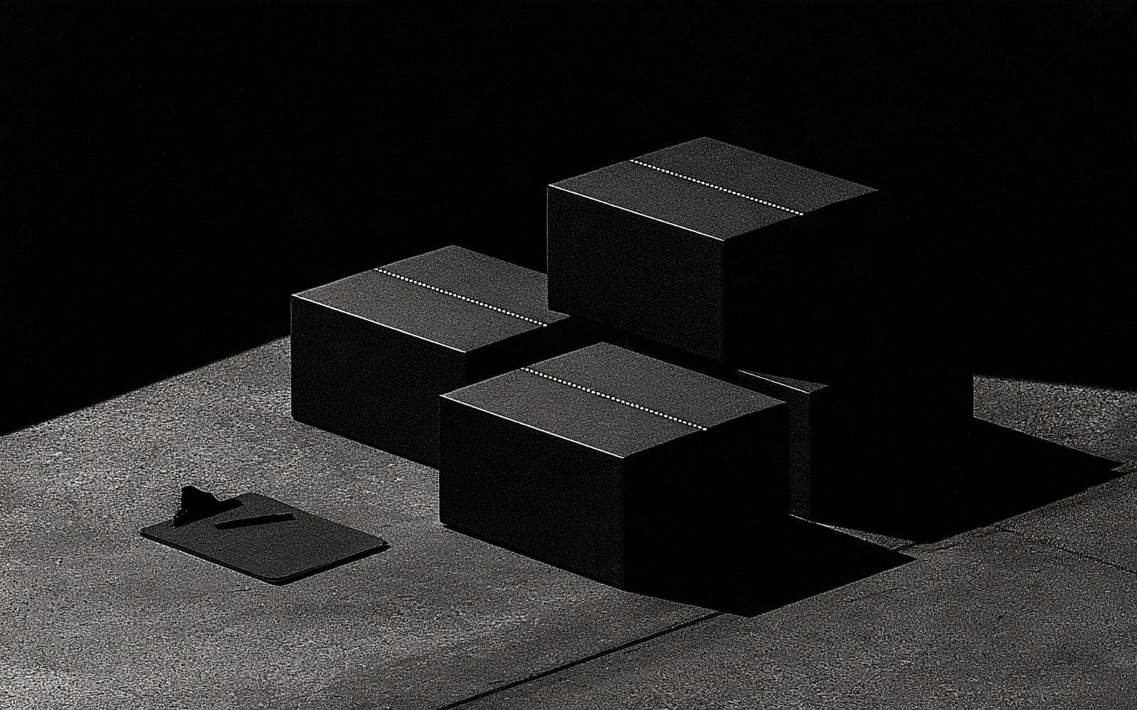 Colis noirs marqués d'une ligne pointillée et un porte-bloc posés au sol, photographie éditoriale sombre en noir et blanc au grain tramé sous une diagonale de lumière.