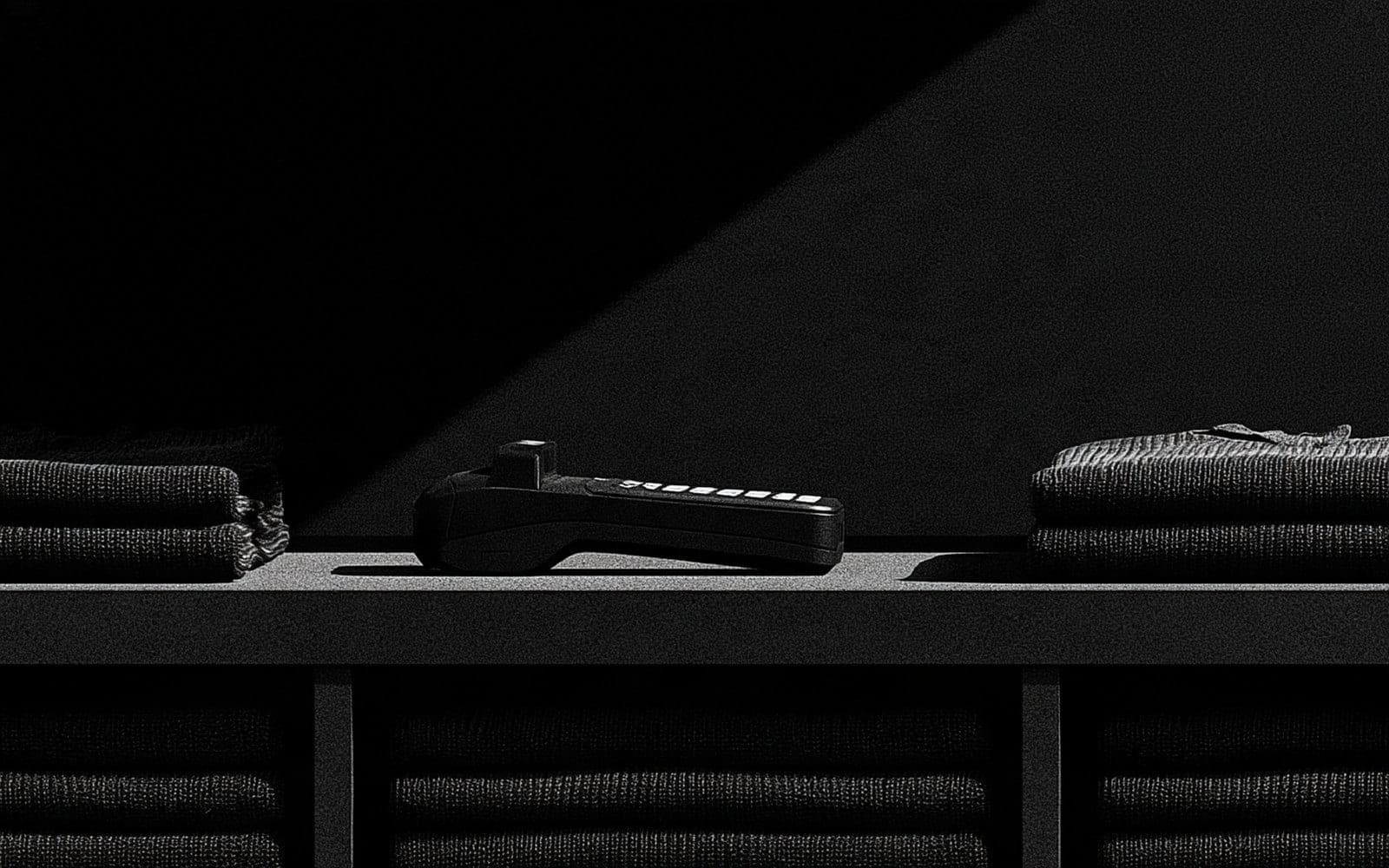 Un terminal de paiement posé sur une étagère entre des piles de vêtements pliés, photographie éditoriale sombre en noir et blanc au grain tramé sous une diagonale de lumière.