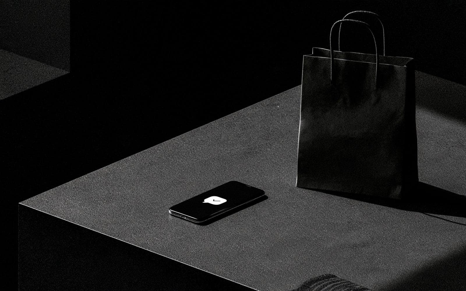 Un smartphone affichant une icône de message validé et un sac de shopping sur une surface sombre, photographie éditoriale en noir et blanc au grain tramé sous une lumière diagonale.