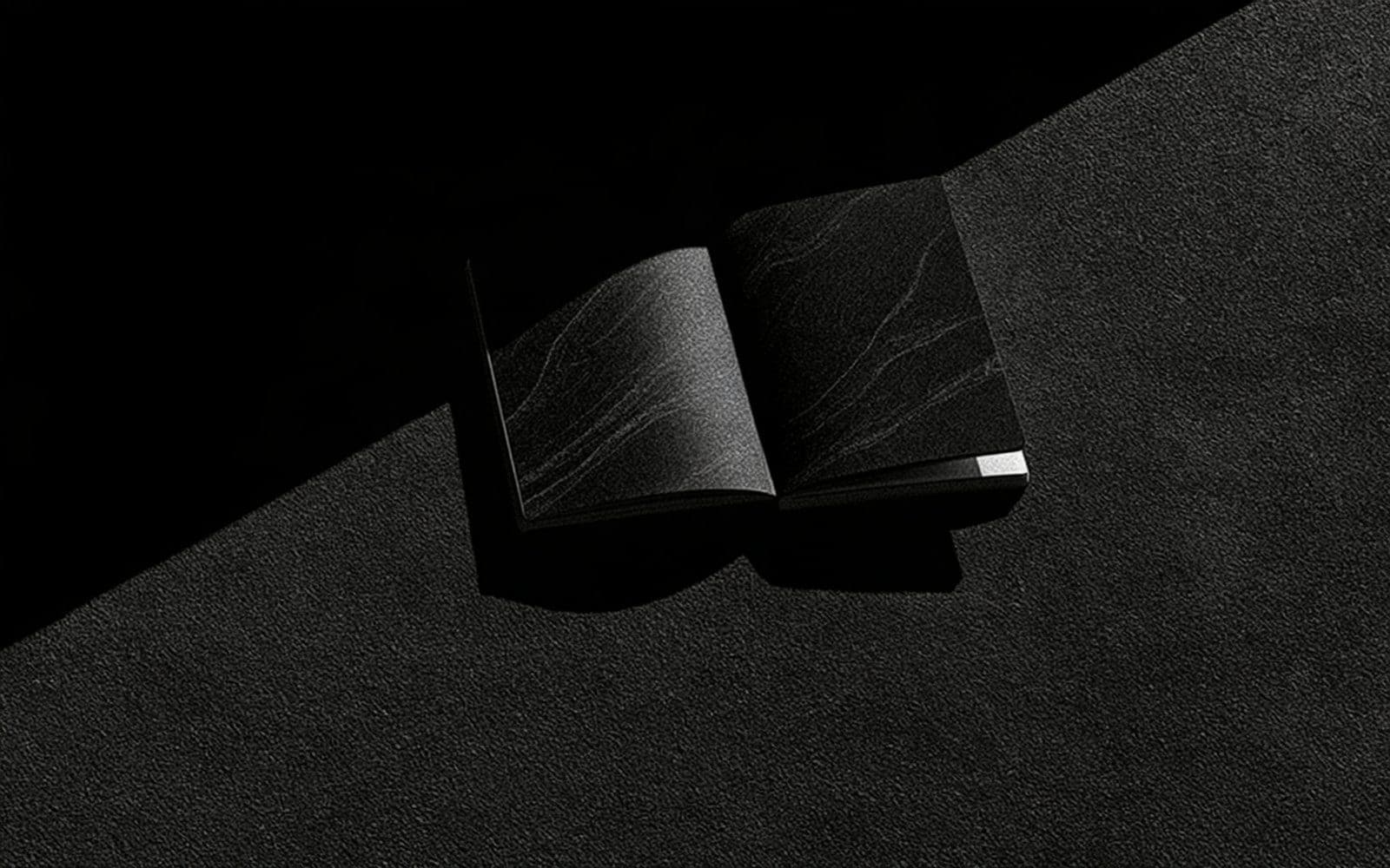 Un carnet ouvert sur une surface granuleuse, photographie éditoriale sombre en noir et blanc au grain tramé sous une diagonale de lumière tranchante.