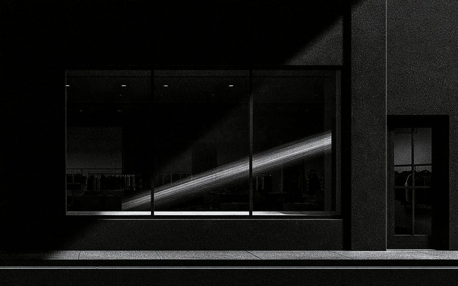 Une vitrine de boutique la nuit avec des silhouettes de vêtements, photographie éditoriale sombre en noir et blanc au grain tramé traversée d'une diagonale de lumière.