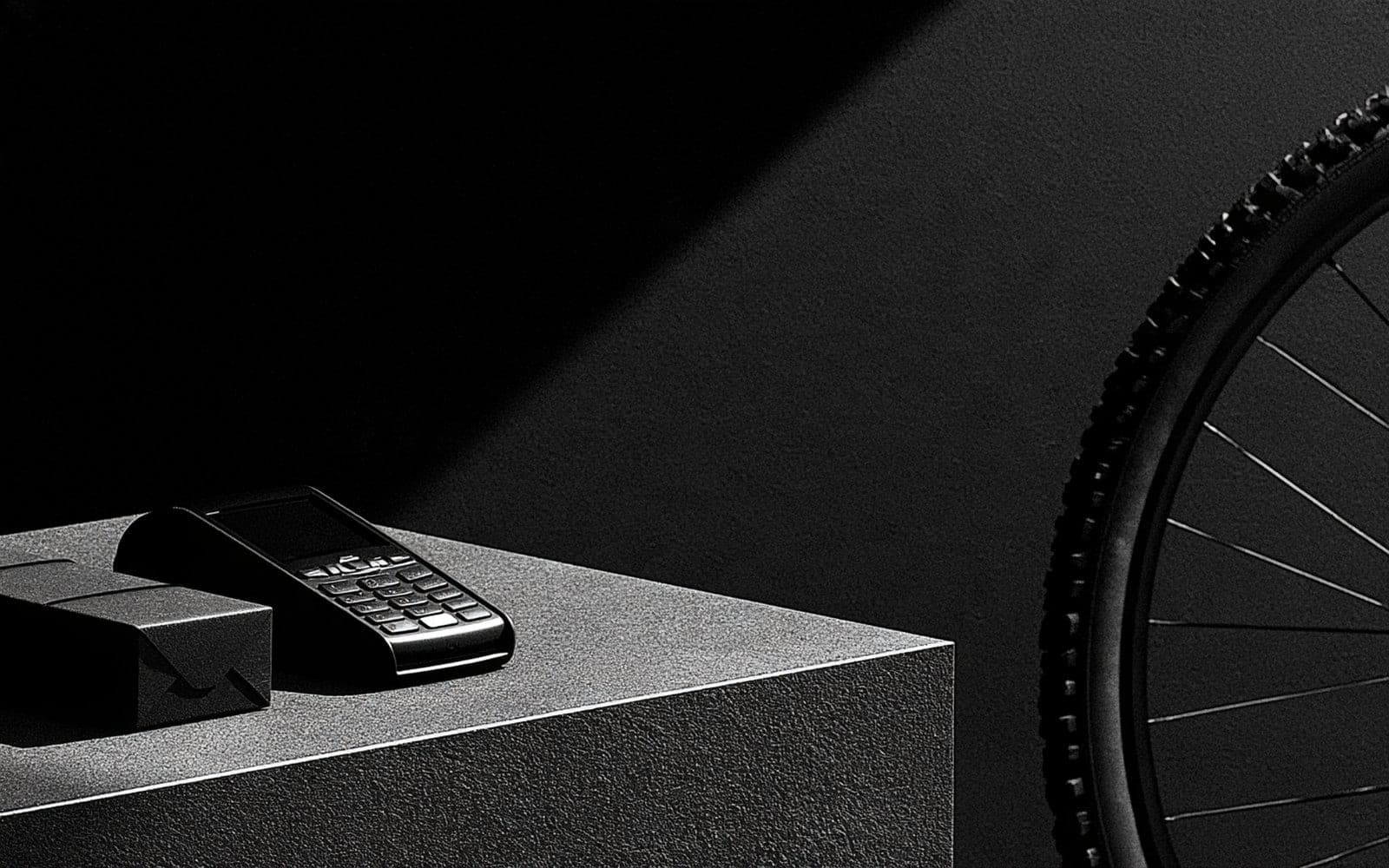 Un terminal de paiement et une boîte cadeau à côté d'une roue de vélo, photographie éditoriale sombre en noir et blanc au grain tramé sous une diagonale d'ombre.