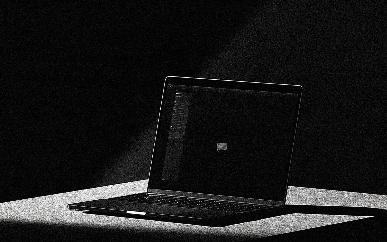 Un ordinateur portable ouvert à l'écran sombre avec une icône de message, photographie éditoriale sombre en noir et blanc au grain tramé sous une diagonale de lumière.