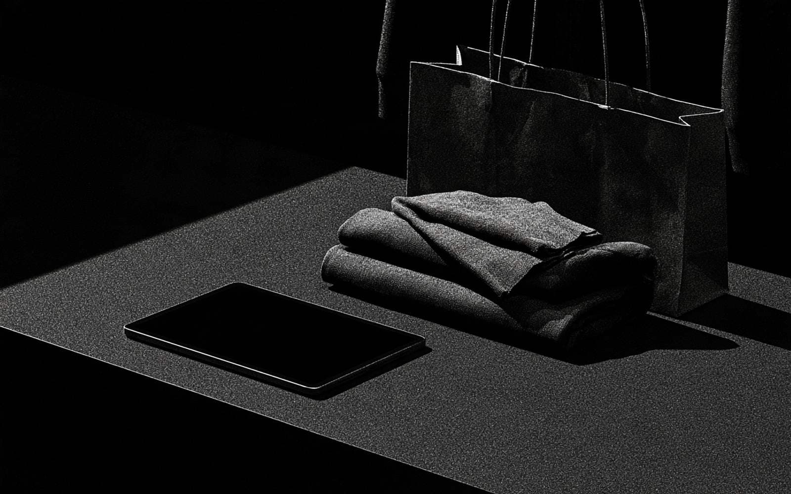 Une tablette de caisse, des vêtements pliés et un sac shopping sur un comptoir sombre, photographie éditoriale en noir et blanc au grain tramé sous une diagonale d'ombre.