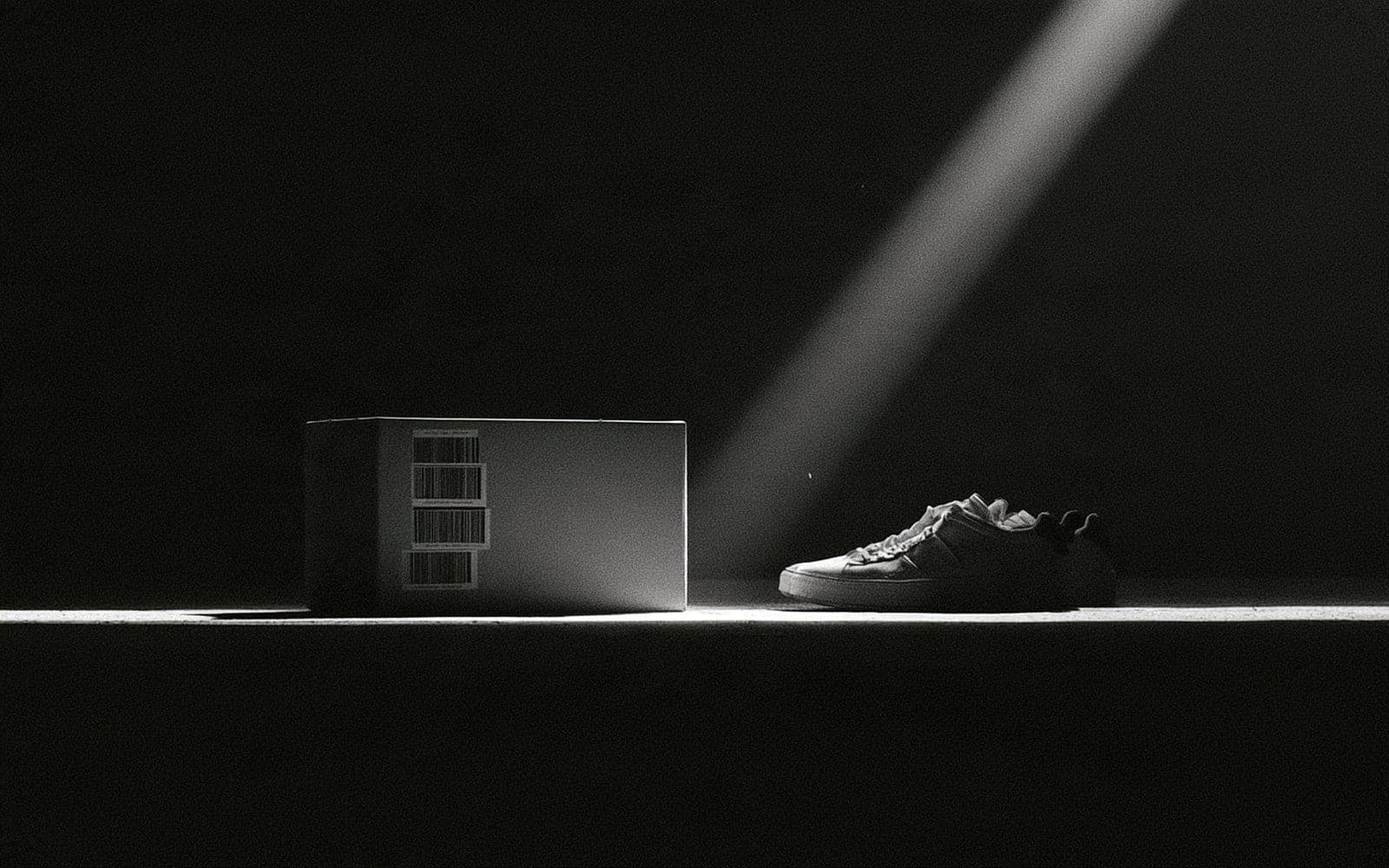 Une boîte d'expédition à codes-barres et une sneaker, photographie éditoriale sombre en noir et blanc au grain tramé sous une diagonale de lumière.
