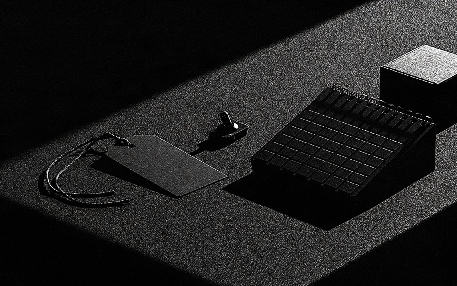 Une étiquette, un interrupteur et un calendrier de bureau sur une surface texturée, photographie éditoriale sombre en noir et blanc au grain tramé sous un éclairage directionnel.