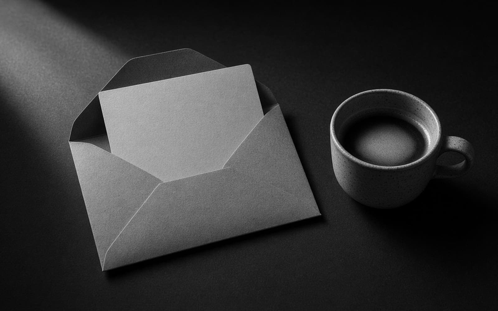 Une enveloppe kraft ouverte et une tasse en céramique sur un bureau sombre, photographie éditoriale en noir et blanc au grain tramé sous une lumière latérale douce.