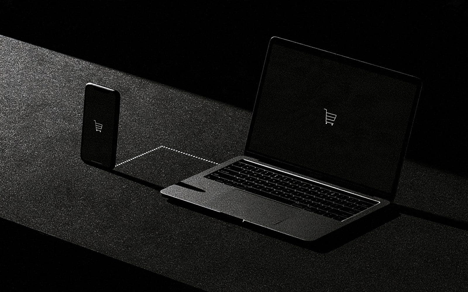 Un smartphone et un ordinateur portable affichant une icône panier reliés par une ligne pointillée, photographie éditoriale sombre en noir et blanc au grain tramé sous une diagonale de lumière.
