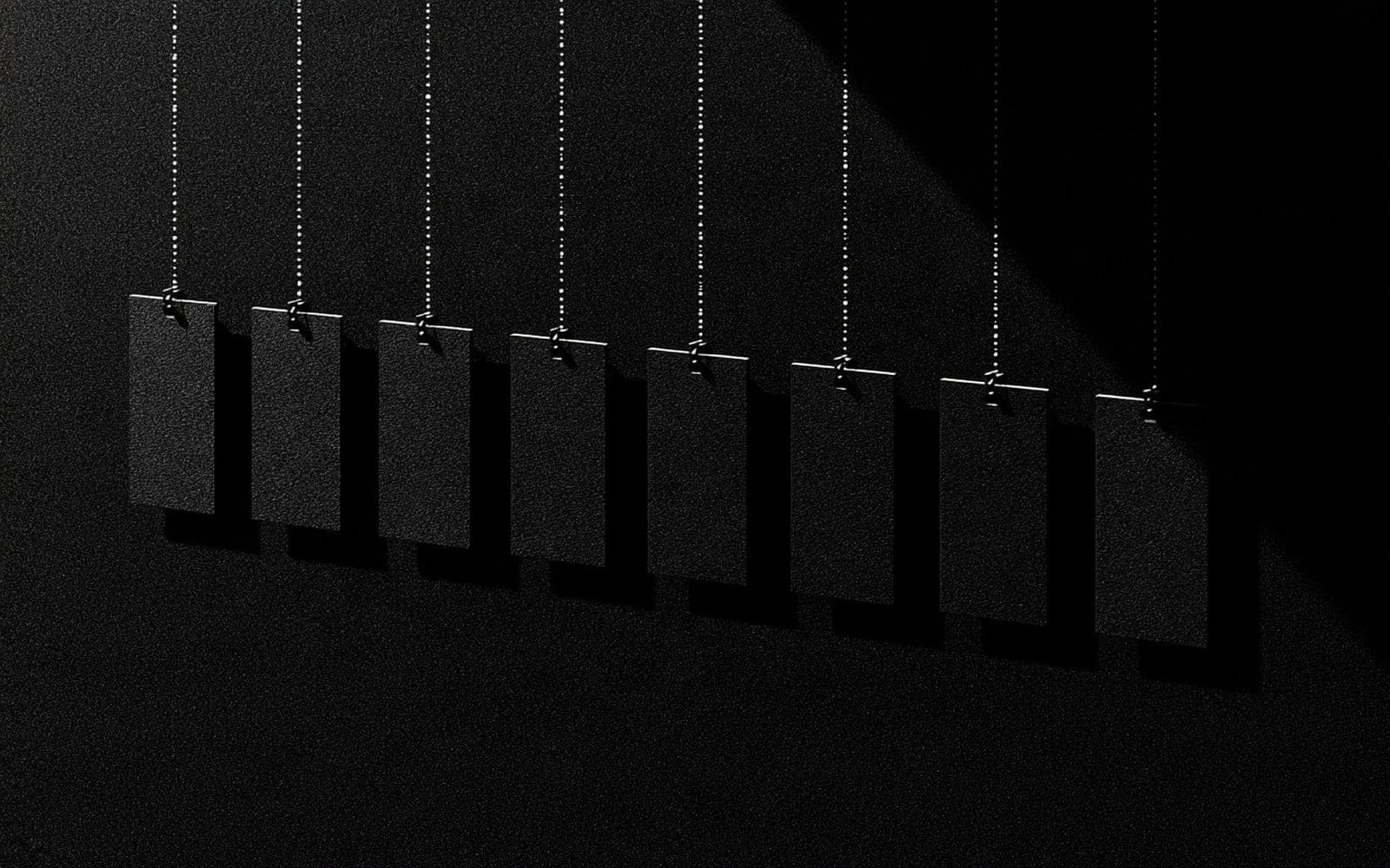 Huit étiquettes noires suspendues à de fines chaînes métalliques, photographie éditoriale sombre en noir et blanc au grain tramé sous une lumière directionnelle.